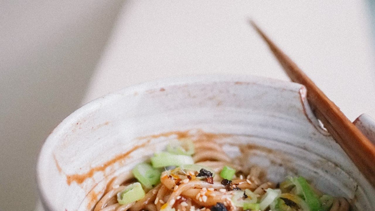 A bowl of spicy peanut noodles garnished with crushed peanuts, sesame seeds, and sliced green onions